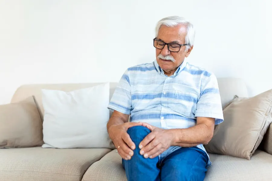Älterer Mann mit Gonarthrose und Knieschmerzen sitzt zu Hause auf dem Sofa und hält sein schmerzendes Knie – Gesundheitsproblem, leidender Senior.